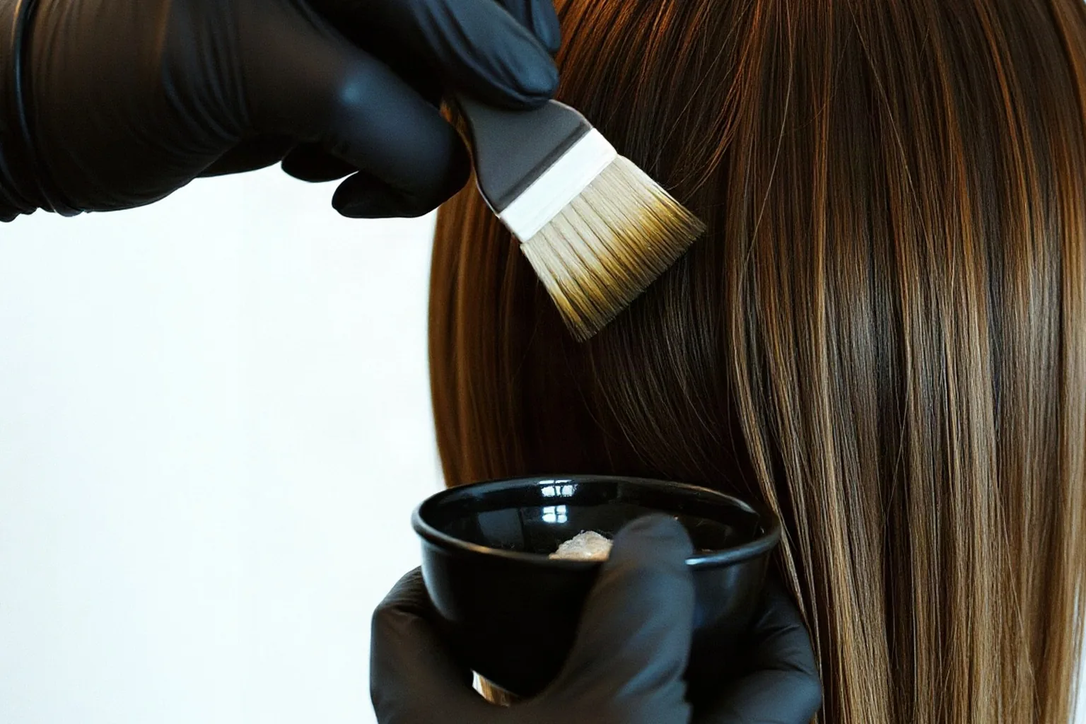 Mani con i guanti stendono il colore con il pennello su una ciocca di capelli castani, con la ciotola del colore