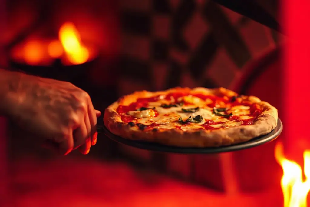 Una pizza appena sfornata sulla pala davanti alla fiamma del forno a legna